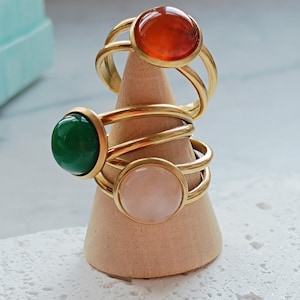 May include: Three gold-colored rings with large, round gemstones in orange, green, and pink are displayed on a wooden cone. The rings have a double-band design. The gemstones are the focal point, showcasing their vibrant colors and polished surfaces.