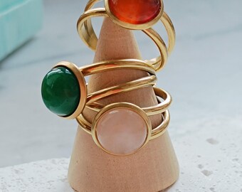 May include: Three gold-colored rings with large, round gemstones in orange, green, and pink are displayed on a wooden cone. The rings have a double-band design. The gemstones are the focal point, showcasing their vibrant colors and polished surfaces.