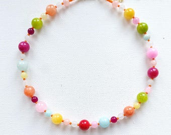 May include: A colorful beaded necklace featuring a variety of round beads in shades of pink, green, yellow, orange, and blue. The necklace has a gold-colored clasp and an orange string.