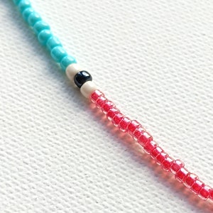 May include: A close-up of a beaded necklace with turquoise, white, and red beads. The necklace has a black bead in the center.
