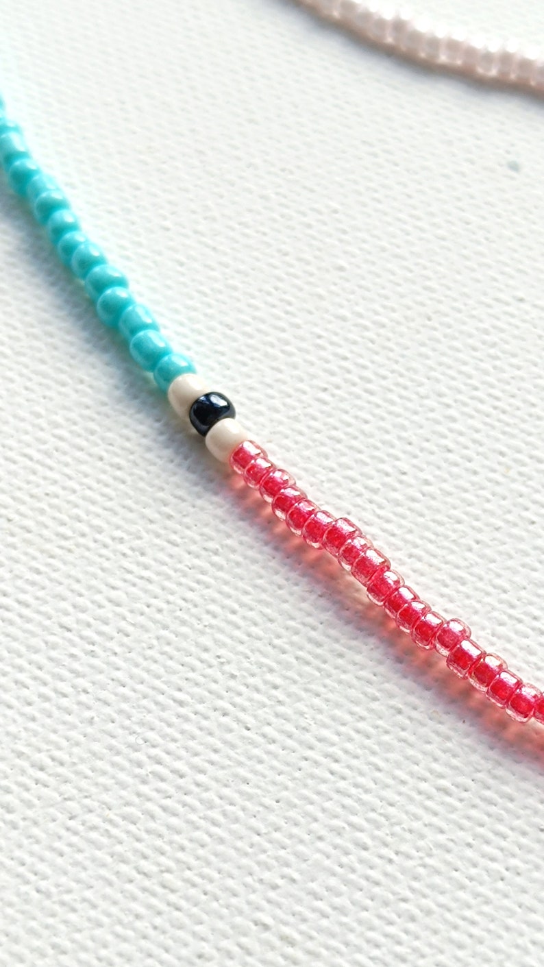May include: A close-up of a beaded necklace with turquoise, white, and red beads. The necklace has a black bead in the center.