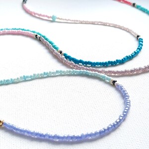 May include: Four layered necklaces with different colored beads. The necklaces are made of small, round beads in shades of blue, pink, and red. The necklaces are layered on top of each other, with the shortest necklace on top and the longest necklace on the bottom.