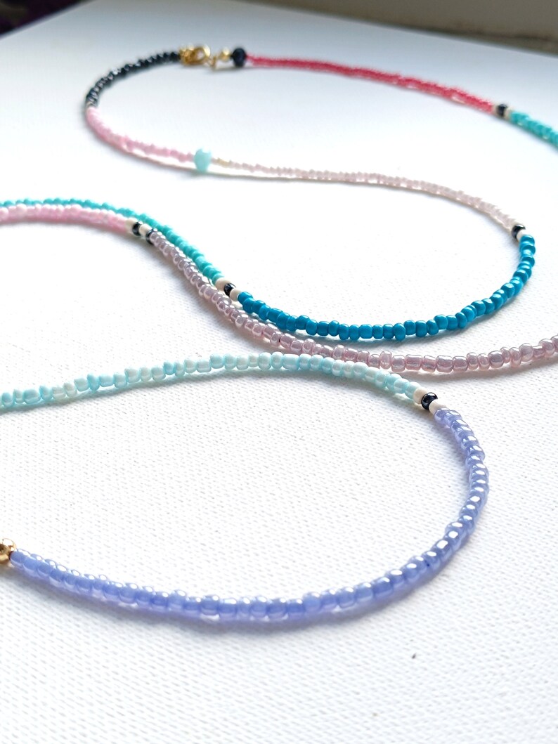 May include: Four layered necklaces with different colored beads. The necklaces are made of small, round beads in shades of blue, pink, and red. The necklaces are layered on top of each other, with the shortest necklace on top and the longest necklace on the bottom.