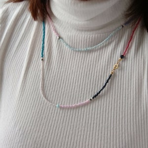 May include: A layered necklace with pink, blue, and black beads. The necklace has a gold clasp and a single turquoise bead.