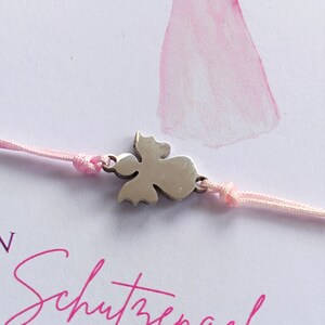 Peut inclure: Un bracelet en cordon rose avec un charm en forme d'ange argent&eacute;. Le fond pr&eacute;sente une illustration &agrave; l'aquarelle d'un ange rose et le texte allemand "Schutzengel F&Uuml;R DICH".