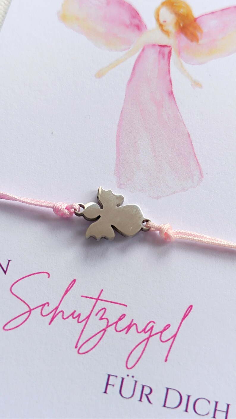 Peut inclure: Un bracelet en cordon rose avec un charm en forme d'ange argent&eacute;. Le fond pr&eacute;sente une illustration &agrave; l'aquarelle d'un ange rose et le texte allemand "Schutzengel F&Uuml;R DICH".