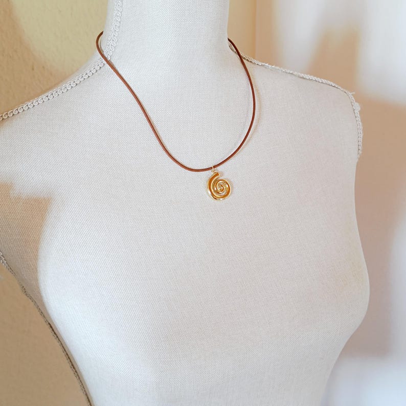 May include: A gold spiral pendant necklace with a brown cord. The pendant features an intricate, open design. The necklace is displayed on a white mannequin, highlighting the jewellery's design and style. The necklace is a simple, elegant accessory.