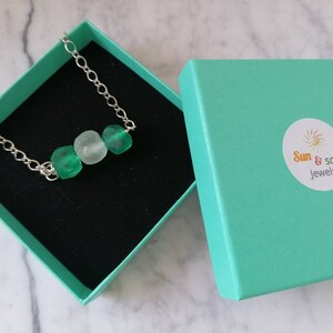 Peut inclure: Un collier en cha&icirc;ne argent&eacute;e avec trois perles en verre vert et blanc. Le collier est dans une bo&icirc;te cadeau turquoise avec le logo "Sun & Soul Jewels".