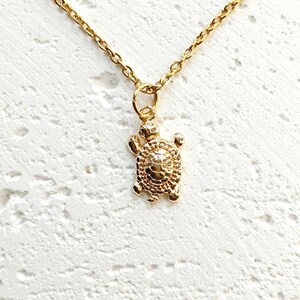 May include: Gold-colored necklace featuring a turtle pendant. The turtle pendant has a textured shell and detailed flippers and head. The chain is delicate and gold-toned, with a lobster clasp. The necklace is displayed against a white textured background.