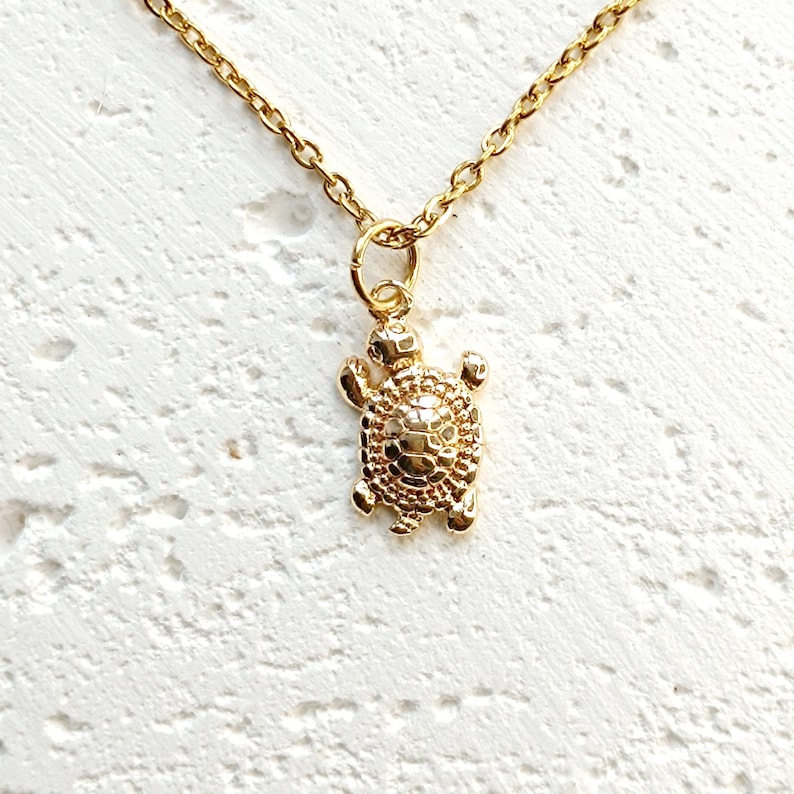 May include: Gold-colored necklace featuring a turtle pendant. The turtle pendant has a textured shell and detailed flippers and head. The chain is delicate and gold-toned, with a lobster clasp. The necklace is displayed against a white textured background.