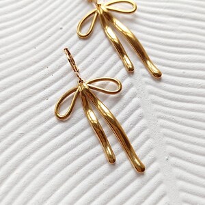 May include: A pair of gold-toned earrings with a unique abstract design. The earrings feature a bow-shaped top and two long, curved strands that dangle below.
