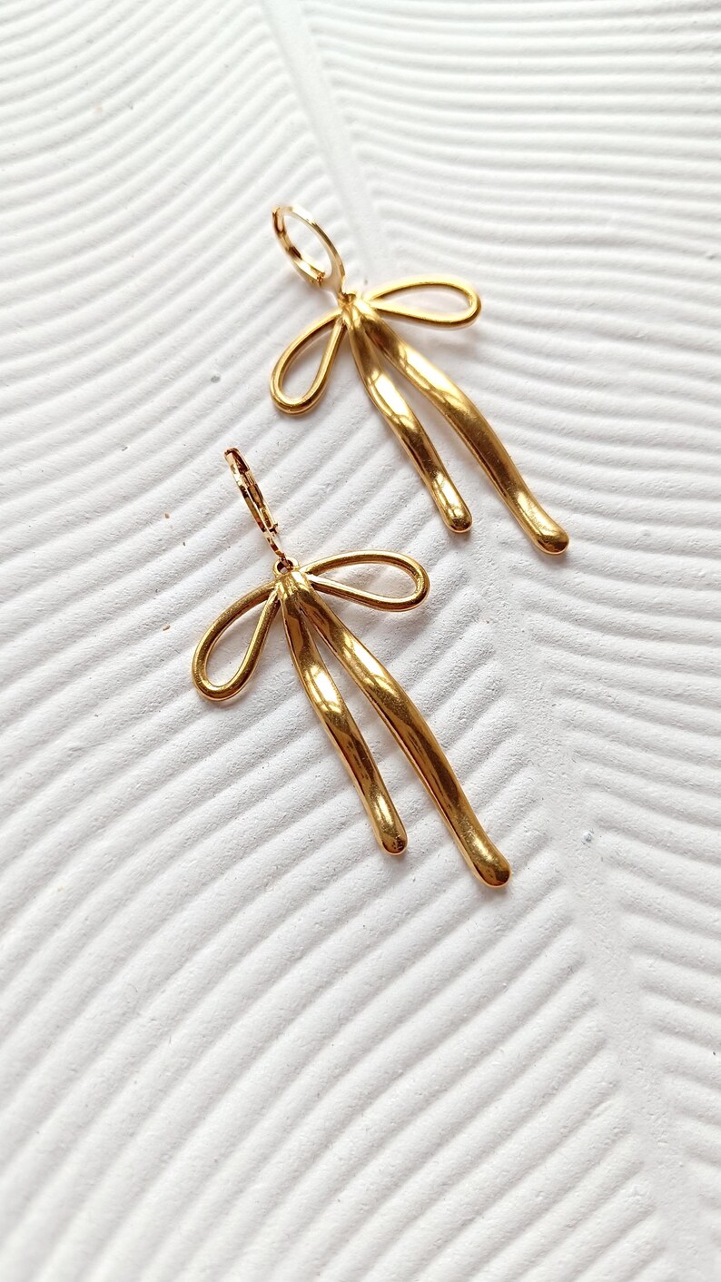 May include: A pair of gold-toned earrings with a unique abstract design. The earrings feature a bow-shaped top and two long, curved strands that dangle below.
