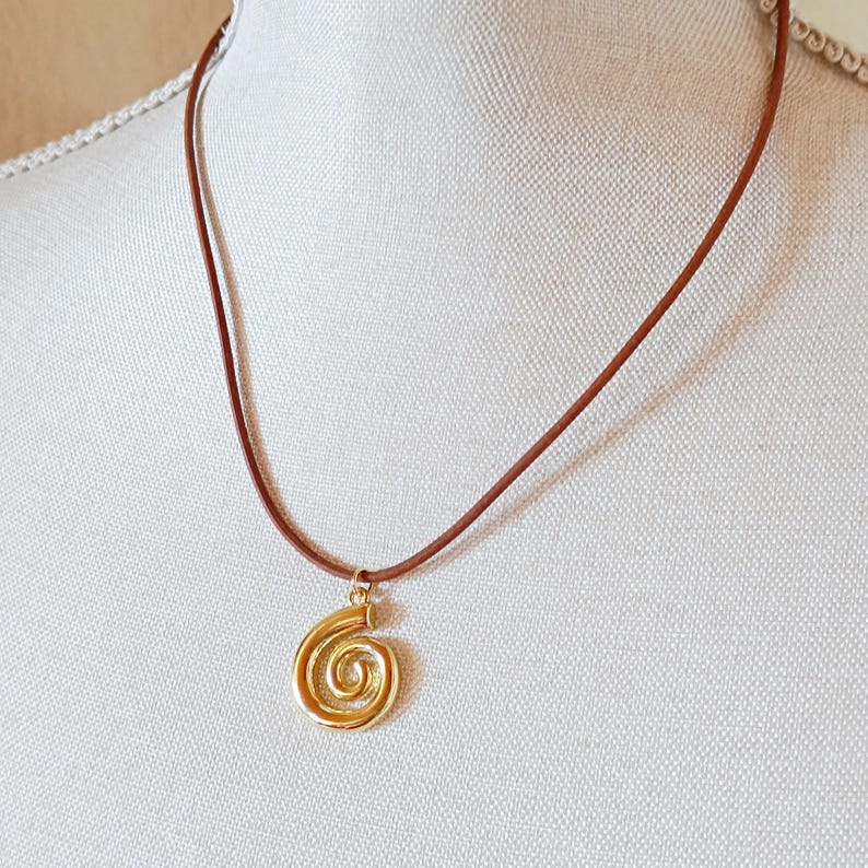 May include: A gold spiral pendant necklace on a brown cord. The pendant is a wire-wrapped design, and the cord is a thin, leather-like material. The necklace is displayed on a mannequin.