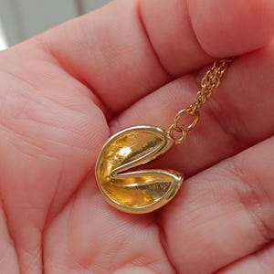 Puede incluir: Un collar con colgante de galleta de la fortuna dorada. El colgante est&aacute; abierto, revelando el interior. La cadena del collar tambi&eacute;n es dorada. El collar se sostiene en la mano de una persona.