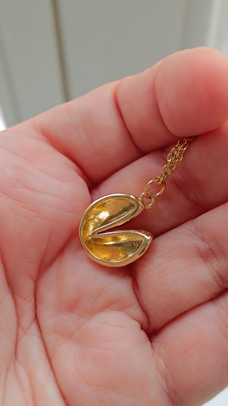Puede incluir: Un collar con colgante de galleta de la fortuna dorada. El colgante est&aacute; abierto, revelando el interior. La cadena del collar tambi&eacute;n es dorada. El collar se sostiene en la mano de una persona.