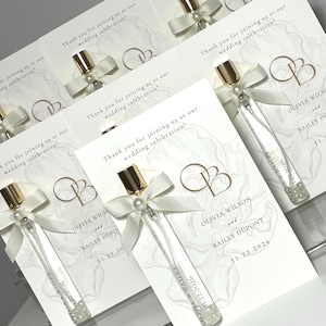 Può includere: Un set di cartoline segnaposto per matrimonio, ognuna con una boccetta di profumo in vetro trasparente con tappo dorato e un fiocco in nastro bianco con perla. Le cartoline riportano il testo 'Thank you for joining us at our wedding celebration!', 'Olivia Wilson and Bailey Dupont' e '11.22.2026' con uno sfondo floreale.