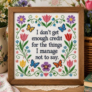 May include: A square cross-stitch embroidery in a wooden frame featuring the black text I don't get enough credit for the things I manage not to say. The design is surrounded by a colourful floral border with tulips, daisies, and butterflies.