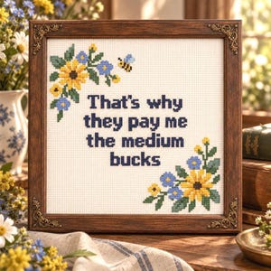 Op de afbeelding: Een vierkant kruissteek borduurwerk in een donkerbruine houten lijst met versierde hoeken. De witte stof bevat de tekst "That's why they pay me the medium bucks" in donkerblauw, omringd door gele en blauwe bloemmotieven en een bij.