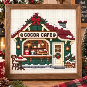 May include: A framed cross-stitch embroidery depicting a winter scene of a building labelled COCOA CAFE. The structure features a dark green exterior, a red roof covered in snow, a red bow, and a white door with a wreath. Mugs and treats are visible inside.