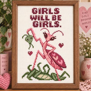 May include: A framed cross-stitch embroidery featuring the text GIRLS WILL BE GIRLS in dark red. The design depicts a pink praying mantis holding a small green insect, surrounded by small red hearts, all on a white fabric background.
