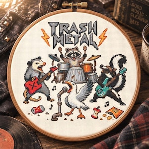 May include: A circular wooden embroidery hoop containing a cross-stitch design of a band of animals playing instruments. The text TRASH METAL is stitched at the top with orange lightning bolts. An opossum, raccoon, seagull, and skunk are depicted.