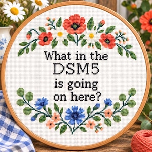 Puede incluir: Un bastidor de bordado circular de madera con tela blanca y un diseño de punto de cruz. El texto dice "What in the DSM5 is going on here?" rodeado por una corona floral de amapolas rojas, margaritas blancas, acianos azules y hojas.