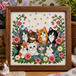 May include: A square cross-stitch embroidery in a brown wooden frame featuring eight cats of various colors and patterns surrounded by pink, blue, and white flowers and green vines, with small yellow stars scattered across the white fabric background.