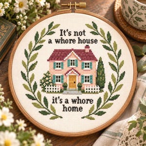 May include: A circular wooden embroidery hoop containing a cross-stitch design of a pink house with a dark red roof, teal shutters, and a yellow door. The text reads: It's not a whore house, it's a whore home. A green leaf wreath surrounds the house.