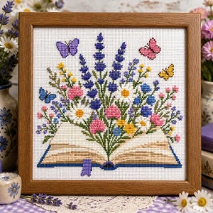 May include: A framed cross-stitch embroidery featuring an open book with a vibrant bouquet of purple lavender, white daisies, pink and yellow wildflowers, and three small butterflies in purple, pink, and yellow, all set against a white grid fabric.