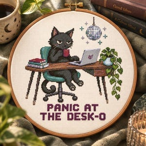 Puede incluir: Un bordado de punto de cruz en un bastidor de madera que muestra un gato negro sentado en un escritorio con un portátil, una taza y libros. Una bola de discoteca cuelga arriba y hay una planta colgante. El texto dice: PANIC AT THE DESK-O.