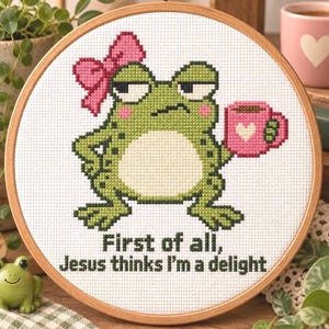 May include: A circular wooden embroidery hoop containing a cross-stitch design of a green frog with a pink bow, holding a pink mug with a white heart. The text below the frog reads, First of all, Jesus thinks I'm a delight.