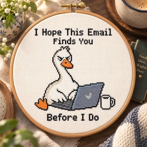 May include: A circular wooden embroidery hoop containing a cross-stitch design on white fabric. The design features a white goose with an orange beak and feet, typing on a blue laptop next to a white mug. Text reads: I Hope This Email Finds You BEFORE I Do.