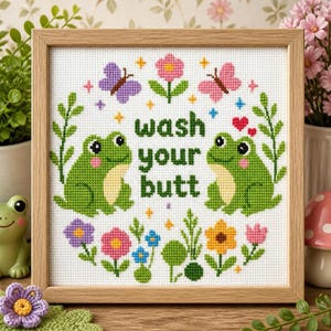 Può includere: Un ricamo a punto croce quadrato in una cornice di legno chiaro raffigurante due rane verdi, fiori colorati, farfalle e il testo "wash your butt" in lettere verdi. Il disegno è centrato su tessuto bianco con un bordo floreale decorativo.