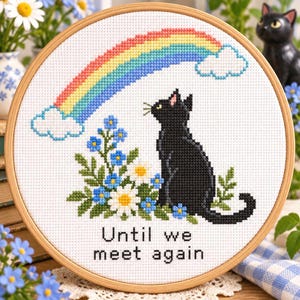 May include: A circular wooden embroidery hoop containing a cross-stitch design of a black cat sitting beside blue and white flowers. Above the cat is a colorful rainbow with white clouds. The text "Until we meet again" is stitched below.