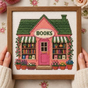 May include: A cross-stitch embroidery in a wooden frame depicting a pink bookstore with a green roof, striped awnings, and shelves filled with colourful books. The word BOOKS is stitched in black letters above a central pink door with potted plants.