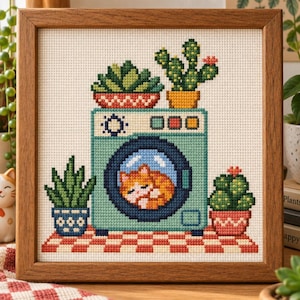 May include: A cross-stitch embroidery in a wooden frame depicting a light blue washing machine on a red and white checkered surface. Inside the circular door, an orange cat is curled up sleeping. Four potted cacti surround the machine.