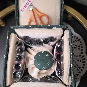May include: A dark green fabric sewing kit box with a white floral pattern, containing orange Singer scissors, multiple spools of thread in clear plastic sleeves, and a central green fabric-covered pincushion with white floral accents.