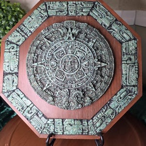 Op de afbeelding: Een achthoekige houten wandplaat met een centraal cirkelvormig reliëf van de Azteekse zonnesteen. Het reliëf heeft een verweerde groen-grijze patina afwerking. De houten lijst is bruin met bijpassende groen-grijze uitgesneden randaccenten.