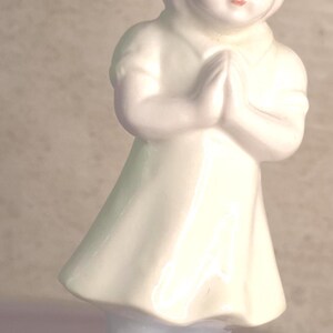 May include: A white porcelain figurine of a child with hands clasped in prayer. The child wears a short-sleeved dress and stands on a small, rectangular base with blue decorative accents and text printed on the front edge of the base.