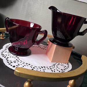 Może przedstawiać: Para naczyń ze szkła w kolorze ciemnej czerwieni, cukiernica z dwoma uchwytami oraz dzbanek na śmietankę z jednym uchwytem, oba ozdobione delikatnym, wytrawianym wzorem kwiatowym na zewnątrz i osadzone na okrągłych podstawach.