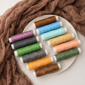 Peut inclure: Une collection de douze bobines de fil cylindriques de différentes couleurs, notamment marron, bleu clair, vert menthe, jaune, pêche, violet, gris foncé, vert vif, anthracite, orange, vert olive et marron foncé, disposées sur une assiette ronde.