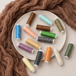 Puede incluir: Una colección de quince pequeños carretes cilíndricos de hilo de coser en varios colores, incluyendo azul, marrón, melocotón, verde, gris, morado, amarillo, naranja y crema, dispuestos sobre un plato blanco redondo con tela marrón texturizada.
