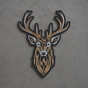 May include: An embroidered patch featuring the head of a deer with large antlers. The design uses shades of tan, brown, and white thread to create a detailed, textured appearance, all outlined with a thick black border against a gray background.