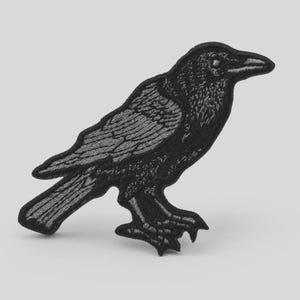 May include: An embroidered patch in the shape of a standing raven. The design features black and dark gray thread detailing to define the bird's feathers, beak, and eye, creating a textured appearance with a clear outline against the background.