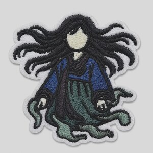 May include: An embroidered patch featuring a figure with long, flowing black hair, a cream-colored face, a blue top, and a dark green skirt. The figure has a ghostly appearance with wispy, trailing edges and is outlined in white thread.