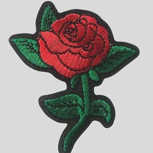 May include: An embroidered patch featuring a vibrant red rose with detailed green leaves and a stem, set against a solid black background. The design is rendered with dense stitching, showcasing the layered petals of the flower and the textured foliage.