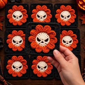 May include: A collection of square crochet motifs featuring a central cream-colored skull surrounded by orange flower petals, all set against a black square background. One motif is held up to display the detailed stitching and color contrast.