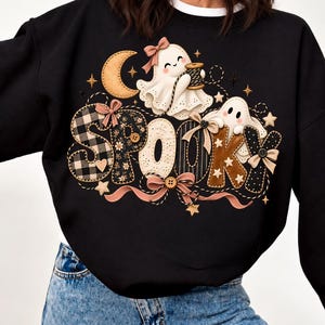 Könnte beinhalten: Ein schwarzes Langarm-Sweatshirt mit einem Grafikdruck des Wortes SPOOKY im Patchwork-Stil mit verschiedenen Mustern, Schleifen und Knöpfen. Zwei weiße Geisterillustrationen befinden sich über dem Text, einer hält eine Garnrolle.