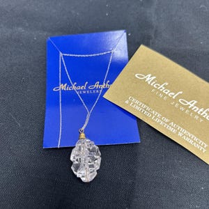 May include: A silver-tone chain necklace with a clear, faceted, irregular-shaped crystal pendant. The necklace is displayed on a blue Michael Anthony Jewelers card next to a gold-coloured certificate of authenticity card with matching text.