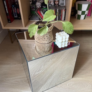 Peut inclure: Une table d'appoint carrée à effet miroir supportant une plante pothos aux feuilles vertes et crème dans un cache-pot en crochet beige et une bougie carrée à bulles blanche. La surface réfléchit la plante et la bougie avec symétrie.